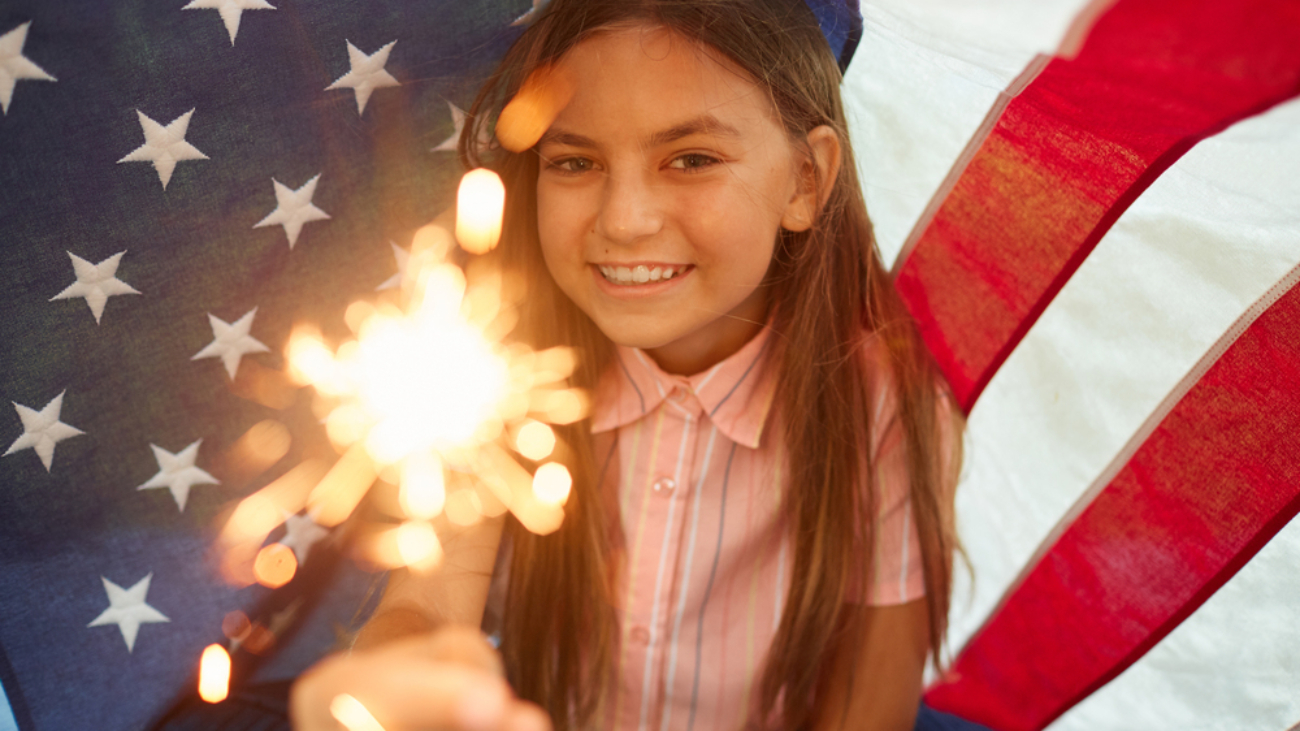 Portrait of cute girl holding sparkling lights smiling at camera against USA flag background, copy space