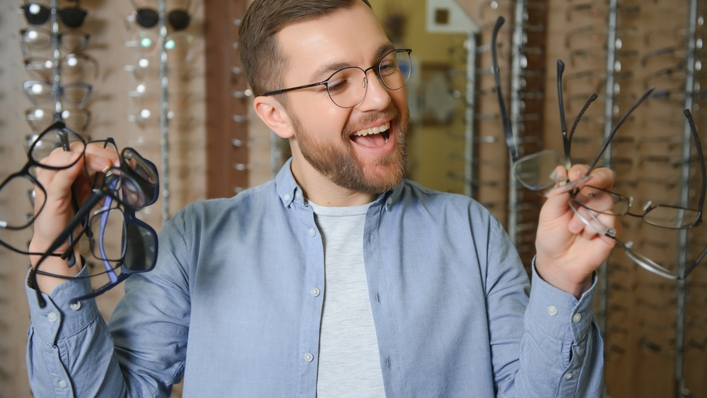 In Optics Shop. Portrait of male client holding and wearing different spectacles, choosing and trying on new glasses at optical store. Man picking frame for vision correction, closeup.
