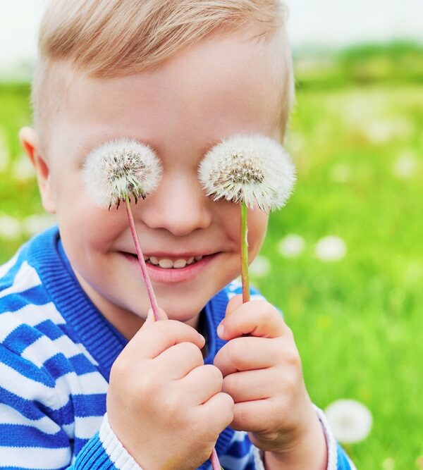Happy face kid smile.Close up portrait joy child outdoors. Little boy playful smiling holding dandelions on eyes as eyeglasses. Joyful childhood, summer day. Background green grass