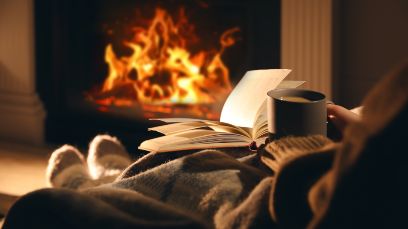 Woman with cup of drink and book near fireplace at home representing a cause of dry eyes in winter