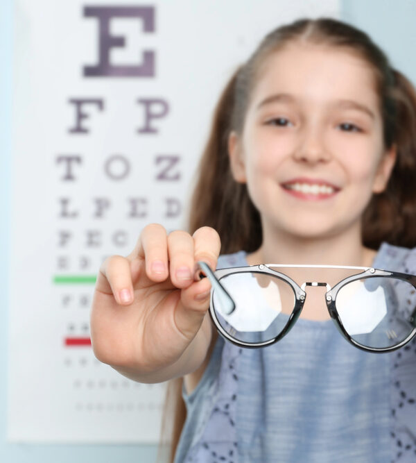 young girl in front of eye chart holding out eye glasses while smiling