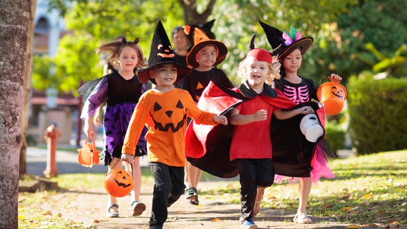 Child in Halloween costume. Mixed race Asian and Caucasian kids and parents trick or treat on street. Little boy and girl with pumpkin lantern and candy bucket. Baby in witch hat. Autumn holiday fun.