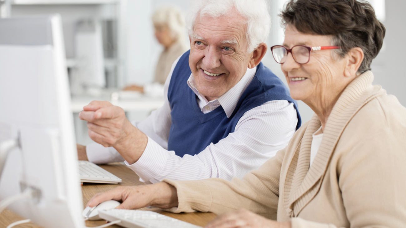 a senior gentleman and woman at a computer keyboard smiling and looking and pointing at the computer screen.