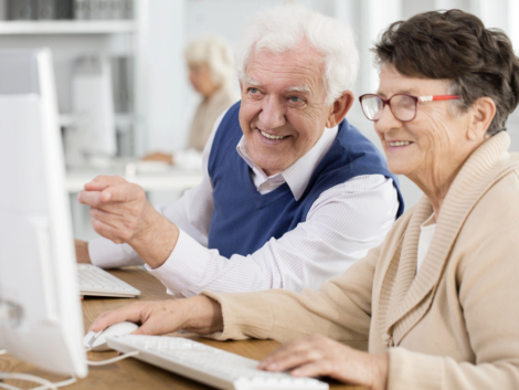 a senior gentleman and woman at a computer keyboard smiling and looking and pointing at the computer screen.
