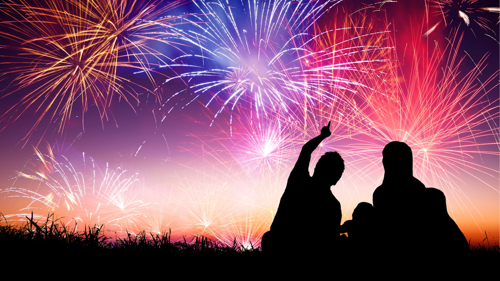 silhouette image of a family sitting in the grass pointing to the sky with fireworks