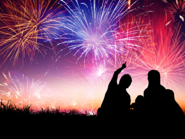 silhouette image of a family sitting in the grass pointing to the sky with fireworks